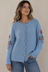 Saco de punto color beige, confeccionado en algodón, con cuello redondo, mangas largas y cierre frontal con botones. Presenta detalles bordados con diseño floral en las mangas y ribetes con rayas azules y blancas en la botonera y el cuello.