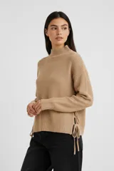 Sweater de tejido de punto color beige, con cuello alto, manga larga y diseño con aberturas laterales ajustables mediante cordones.