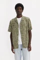 Camisa de manga corta con cuello tipo campamento, confeccionada en tela estampada con diseño de hojas en tonos verde y blanco. Presenta cierre frontal con botones y un bolsillo en el pecho con etiqueta roja distintiva.