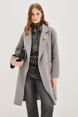 Tapado de paño en color gris, con diseño de corte recto, solapas clásicas, cierre frontal con botones y bolsillos laterales. Presenta un pin decorativo en forma de corazón dorado en la solapa.