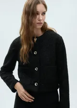 Chaqueta de tejido bouclé en color negro con textura y detalles brillantes. Presenta un diseño recto, cuello redondo, manga larga, cierre frontal con botones efecto joya y dos bolsillos de parche delanteros.