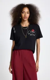 Remera negra de algodón con cuello redondo y mangas cortas. Presenta un bordado frontal con la frase 'L'amour est déclaré' acompañada de un diseño de labios rojos, y un detalle bordado adicional en la manga derecha.