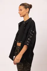 Camisa negra de microfibra con corte oversized. Presenta mangas con detalle de ojalillos y acordonado lateral, doble bolsillo frontal con solapa y botones metálicos. La parte trasera incluye diseño con tachas, ojalillos y cadena.