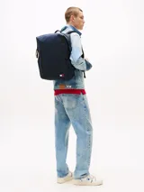 Mochila azul marino con acabado liso, compartimento principal con cierre de cremallera, bolsillo frontal vertical con cremallera, dos bolsillos laterales para botellas y correas ajustables. Incluye parche engomado con el logo de Tommy Jeans en la parte delantera.