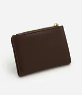 Billetera mediana color marrón, confeccionada en poliuretano con textura lisa y detalle de gamuza en la parte superior. Presenta cierre superior con cremallera dorada y aplicación de tachas metálicas decorativas en el frente.