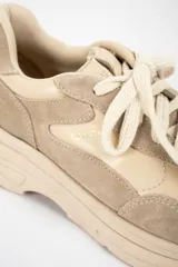 Zapatilla urbana de diseño chunky, confeccionada en una combinación de cuero liso y gamuza en tonos beige y arena. Presenta cordones al tono, suela de plataforma con relieve y detalle de marca grabado en el talón.