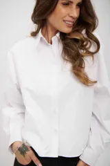 Camisa blanca de corte recto y largo cropped, con cuello clásico, cierre frontal de botones y mangas largas con puños abotonados. Presenta un sutil estampado de pequeños puntos distribuidos por toda la prenda.