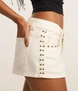 Short de sarga color blanco roto, modelo micro, con trabillas en la cintura, cierre tradicional, bolsillos y tachuelas aplicadas desde los bolsillos hasta los laterales. Presenta costura aparente en contraste.