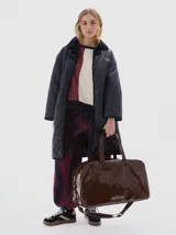 Bolso de fin de semana tipo duffle, confeccionado en material sintético efecto charol color marrón oscuro. Presenta doble asa de mano, correa ajustable extraíble, cierre superior y logo de la marca estampado en el frente.