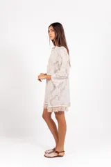Vestido camisero corto de encaje blanco con diseño ornamental, escote en V con cordón ajustable y ojales metálicos, mangas 3/4 acampanadas y ruedo con detalle de flecos de caracoles tejidos a mano.
