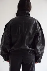 Chaqueta de cuero sintético color negro, con diseño de corte oversize, cuello alto con cierre de botones a presión, hombreras marcadas y ruedo elástico.
