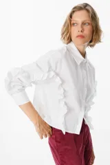 Camisa de corte cropped en color azul, con cuello clásico y cierre frontal de botones. Presenta volados decorativos que recorren la parte frontal y las mangas largas, terminando en puños abotonados.