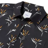 Camisa de manga corta con estampado de flores de ave del paraíso en tonos naranja y blanco sobre fondo negro. Tiene cuello clásico, cierre con botones y bolsillo en el pecho.