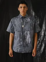 Camisa de manga corta con estampado abstracto en tonos grises, cuello clásico, bolsillos frontales aplicados y cierre frontal de botones a presión.