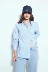 Camisa de silueta amplia y caída fluida, color celeste con rayas verticales blancas. Presenta cuello camisero clásico, cierre frontal con botones y un bolsillo aplicado en el pecho. Las costuras están resaltadas en azul oscuro.