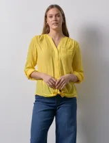 Blusa blanca de gasa marca Zac & Rachel, con cuello a la base, escote en V, abertura central con botones, bolsillo aplicado en el pecho y mangas largas regulables con presilla y botón. Tiene terminación opcional de nudo en el ruedo.