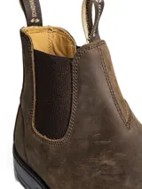 Bota tipo Chelsea de cuero color gris oscuro, con elásticos laterales negros y suela de goma dentada. Presenta tiradores en la parte superior para facilitar el calce.