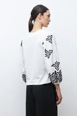 Remera de color crudo con mangas largas abullonadas, decoradas con bordados en color negro. Presenta un diseño de cuello redondo y corte recto.