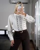 Camisa de manga larga color blanco con bordados geométricos en negro a lo largo de la botonadura frontal y en los puños. Presenta cuello mao y cierre frontal con botones.