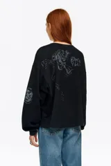 Sudadera negra de corte holgado y manga larga abullonada, con cuello redondo. Presenta detalles de pedrería brillante y estampados sutiles distribuidos por la prenda.