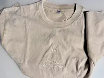 Remera de algodón color beige con efecto acid wash, corte oversize y mangas cortas. Presenta un delicado bordado frontal con la letra M y el texto "MOOD JUST HAPPY".