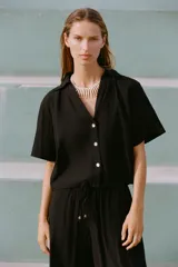 Camisa negra de cuello solapa con escote pico y manga corta. Detalle de nido de abeja en espalda. Cierre frontal con botones.