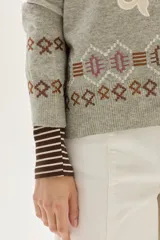 Polera de tejido de punto en color gris, con cuello alto y diseño holgado. Presenta un aplique central en color crema con motivos ornamentales y detalles bordados geométricos en la parte inferior y en las mangas.