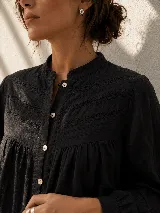Camisa negra de calce amplio y fluido, con cuello mao y cierre frontal de botones. Presenta un canesú con detalles de encaje calado en la parte delantera y mangas largas.