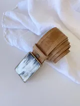 Cinto de cuero color marrón con forro de cuero y hebilla rectangular con diseño de guampa en blanco y negro con marco metálico plateado.