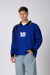 Camiseta de tejido de punto con cuello polo, manga larga y diseño inspirado en camisetas de fútbol retro. Presenta un color azul vibrante con un patrón geométrico lateral en negro y el número 10 estampado en el frente.