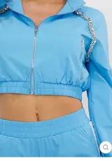 Conjunto deportivo de dos piezas color celeste con franjas blancas en los laterales. Incluye una campera crop de manga larga con cierre frontal, bolsillos y detalles de pedrería brillante en los hombros, y un short a juego con cintura elástica y bolsillos.
