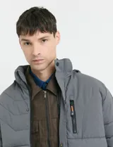 Campera puffer gris oscuro con capucha desmontable, cierre frontal con cremallera y botones a presión, bolsillos laterales con cierre y detalles en color naranja.