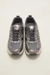 Zapatilla deportiva de estilo urbano en color blanco con detalles metalizados plateados y acentos en negro. Presenta una combinación de malla transpirable y paneles sintéticos, con suela robusta y cordones al tono.