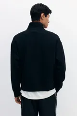 Buzo negro de tejido pesado con interior cepillado suave, cuello elevado con medio cierre y hombros ligeramente caídos. Presenta un estampado frontal con el número "21" en color beige. Puños y bajo en punto acanalado.