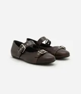 Zapatilla tipo bailarina color marrón oscuro, confeccionada en poliuretano con acabado mate. Presenta punta redondeada, suela plana y dos tiras ajustables con hebillas metálicas plateadas y ojales decorativos sobre el empeine.