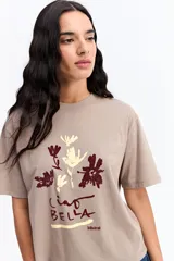 Remera de corte oversize color beige, con cuello redondo y mangas cortas. Presenta un estampado frontal con motivos florales abstractos y la frase 'Ciao Bella' en tonos bordó y crema.