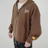 Canguro color marrón con capucha, cierre frontal parcial, bolsillo canguro y cordón ajustable en el dobladillo. Tiene un estampado blanco en el pecho con el texto "PULAU DESIGN DEPT." y un parche amarillo en la manga izquierda.