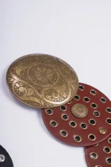 Cinturón de cuero con diseño de discos circulares superpuestos, adornados con tachas y hebillas redondas grabadas. Disponible en negro, marrón y con hebillas en tonos dorado y plateado.