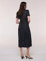 Vestido midi negro con estampado de lunares blancos, de la marca Sharagano. Presenta cuello con solapa, escote en V, cierre frontal con botones y mangas cortas. Tiene un corte en la cintura con lazo ajustable y falda plisada.
