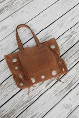 Bolso estilo shopper de gamuza color marrón, con diseño artesanal que incluye flecos laterales y apliques de tachas metálicas circulares. Cuenta con doble asa de hombro y costuras a la vista.