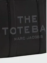 Cartera tipo tote de cuero granulado color tostado, con diseño estructurado y rectangular. Presenta doble asa superior fija y una correa larga ajustable y extraíble. En el frente, destaca el logo estampado con la inscripción 'THE TOTE BAG MARC JACOBS'.