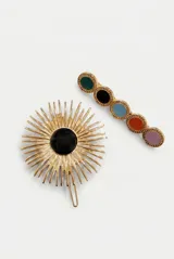 Set de dos hebillas para el pelo con acabado metálico dorado. Incluye una hebilla en forma de sol con centro de piedra negra y una hebilla alargada con detalles circulares de colores.