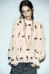 Blusa de cuello subido y manga larga con puños acampanados elásticos. Confeccionada en tejido semitransparente color beige con bordado de lentejuelas oscuras. Presenta cierre en la espalda con abertura y botones ocultos.