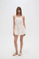 Vestido corto de algodón color blanco, con breteles finos tipo lencero, escote recto con detalle de volado y cintura con elástico fruncido. La falda presenta un diseño holgado con volado en el ruedo.