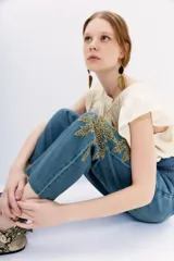 Pantalón de denim azul de tiro alto con cintura estilo paper bag y lazo a tono. Presenta un bordado elaborado con lentejuelas y piedras en forma de palmeras en la parte lateral de la pierna.