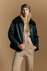 Campera tipo gamulán de cuero sintético negro, con interior y detalles de corderito sintético al tono en cuello y ruedo. Presenta cierre frontal metálico y diseño de silueta relajada.
