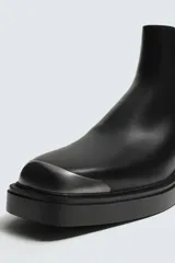 Bota tipo Chelsea de cuero negro con paneles elásticos laterales, suela de goma con volumen y detalle de pieza metálica en la puntera.