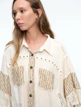 Camisa de corte oversize en color crudo, confeccionada con una mezcla de texturas que incluye paneles de encaje, detalles bordados y aplicaciones de tachas metálicas. Presenta mangas amplias tipo murciélago, cuello clásico y cierre frontal con botones.