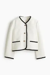 Chaqueta corta en tejido bouclé de color blanco marfil, con ribetes negros en contraste en el cuello en V, la abotonadura, los bolsillos frontales ribeteados, el ruedo y los puños. Presenta botones metálicos dorados con diseño de nudo y está completamente forrada.