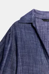 Camisa azul confeccionada en hilatura con 15% de lino. Tiene cuello solapa, escote pico, mangas 3/4 abullonadas, bajo recto y cierre frontal con botones.